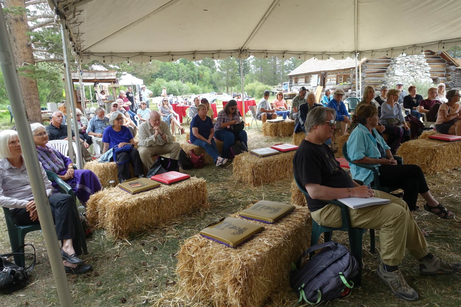 Picture of book festival attendees holding books and sitting on haystacks under a canopy.